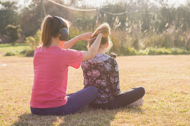 Sister wearing headphone tying her sisters hair in the park