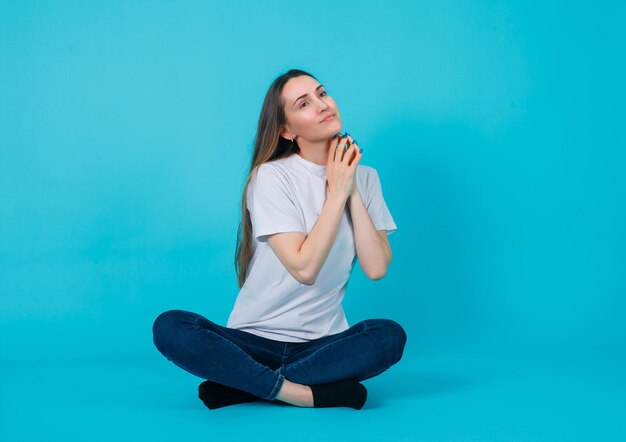 Sitting on floor girl is looking up by holding hands on chest on blue background