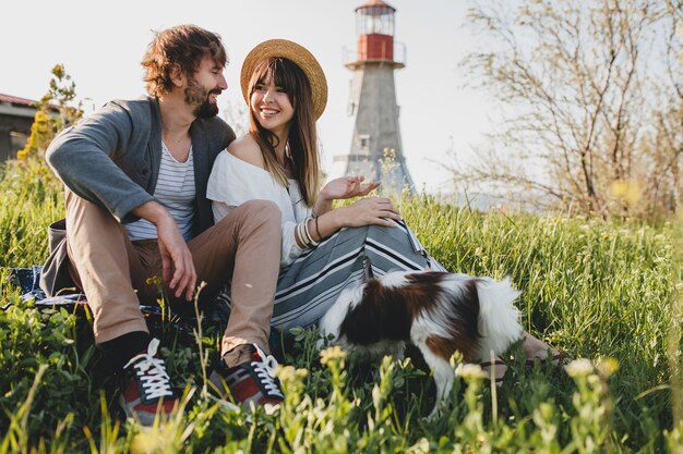 Sitting in grass young stylish hipster couple in love walking with dog in countryside, summer style boho fashion, romantic