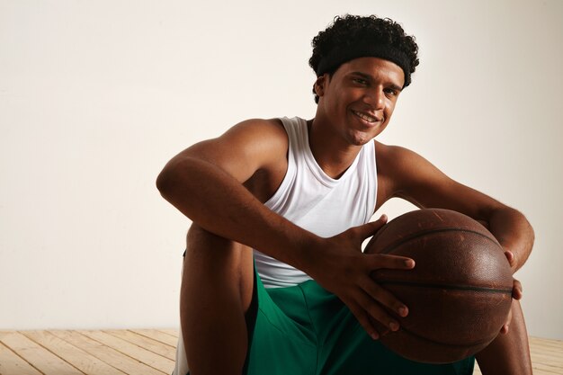 sitting smiling friendly African American basketball player with an afro wearing white and green uniform holding a brown leather ball