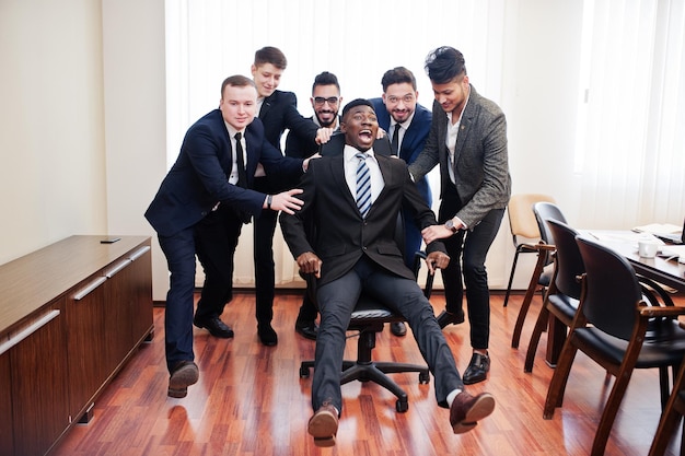 Six multiracial business mans standing at office and roll man on chair Diverse group of male employees in formal wear having fun