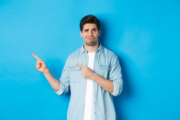 Skeptical adult man pointing fingers left, looking doubtful and unsure, standing over blue background