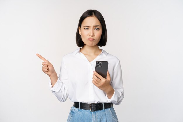 Skeptical asian modern girl holding smartphone pointing finger left at empty space and frowing hesitant has doubts standing over white background