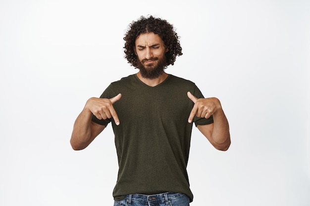 Free photo skeptical bearded man pointing fingers and looking down frowning and grimacing disappointed dislike something standing over white background