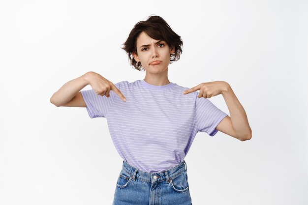 Skeptical brunette girl pointing fingers at product, showing logo sale banner with upset, disappointed face, frowning and sulking unsure, standing over white background