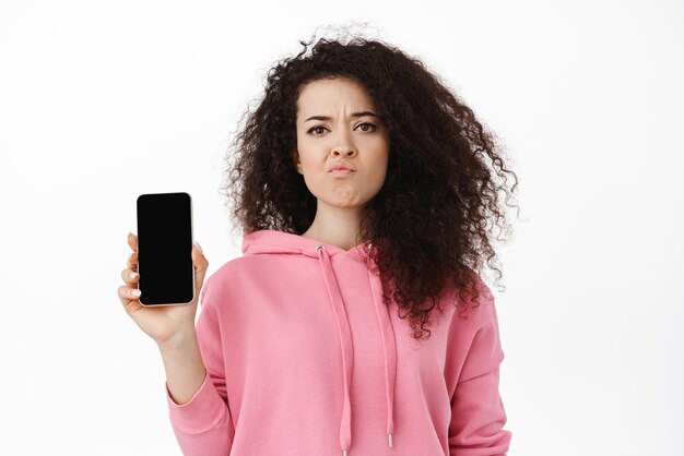 Skeptical brunette woman complaining showing empty mobile screen and grimacing upset disappointed by smartphone app standing in pink hoodie against white background