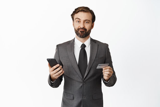 Skeptical businessman in suit grimacing disappointed holding credit card and mobile phone standing over white background