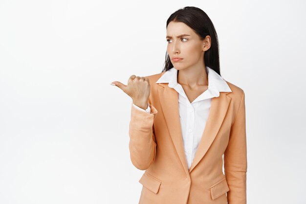 Skeptical businesswoman pointing looking left with frowning face standing in suit against white background