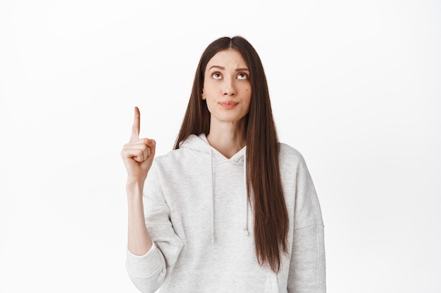 Skeptical caucasian girl with long hair, frowning and having doubts about product, pointing at empty space on top, showing advertisement up, standing against white background