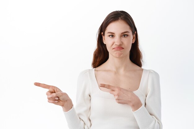 Skeptical and disappointed young woman complaining on bad promotional text, pointing fingers aside at logo banner, grimacing displeased, standing over white wall