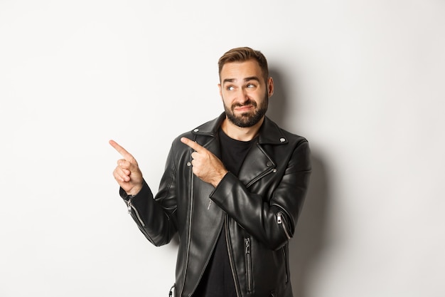 Skeptical and doubtful guy in black leather jacket, shrugging while pointing at upper left corner promo offer, standing