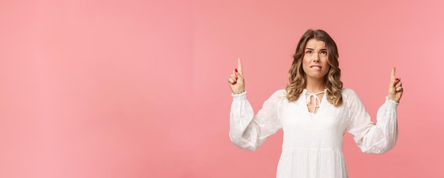 Free photo skeptical and embarrassed blond caucasian woman in white cute dress grimacing cringe from seeing something disgusting frowning doubtful and pointing fingers up strange thing pink background