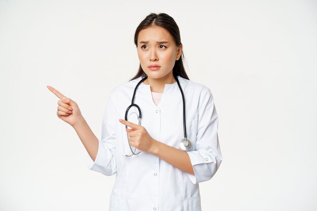 Skeptical female doctor concerned nurse pointing and looking left with worried face expression stand...