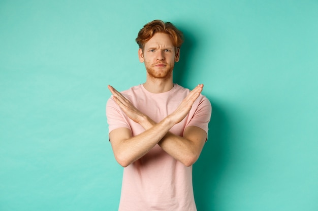 Skeptical redhead man in t-shirt saying no, cross arms on chest and frowning disappointed, prohibit something bad, standing over turquoise background.