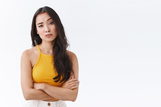 Skeptical, unimpressed pretty east-asian female in yellow top, cross hands on chest defensive and unamused pose, raise one eyebrow and stare judgemental being uninterseted and doubtful