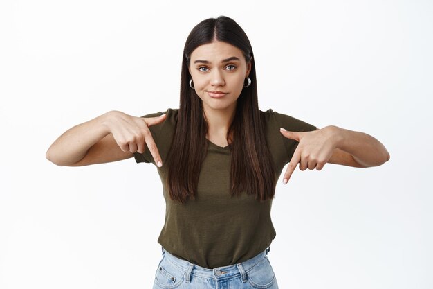 Skeptical young brunette woman having doubts, pointing fingers down at something bad or displeasing, standing against white wall