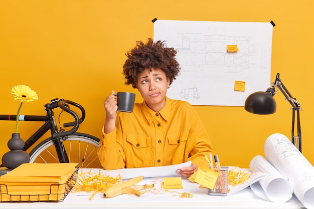 Skilled professional female engineer drinks coffee prepares drawings for house development project being deep in thoughts poses at desktop with papers around 