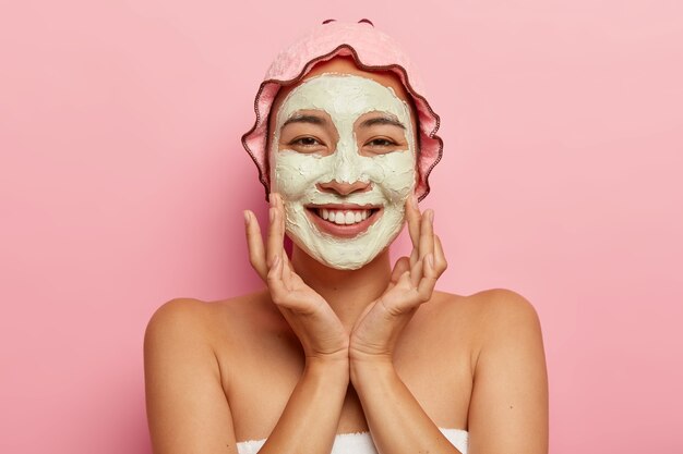 Skin care for all ages. Happy Asian lady with peeling clay mask on face, has beauty treatments, looks pleasantly, touches cheeks, wears shower cap