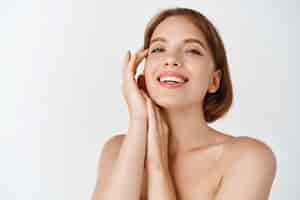 Free photo skin care beauty. smiling natural woman with naked shoulders and healthy clean and fresh skin, looking happy, touching cheek. girl apply facial cosmetics, white wall