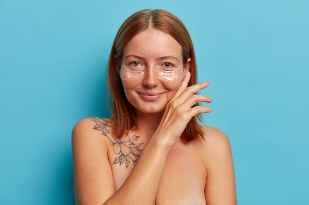 Free Photo skin care and cosmetology procedure. satisfied freckled woman touches face gently, wears hydrogel eye patches, stands naked, has perfect well cared body with charming smile.