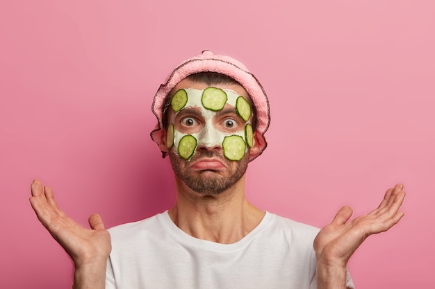 Free photo skin care for men. embarrassed puzzled man applies facial mask with cucumber slices, cleans acnes and pimples, spreads palms