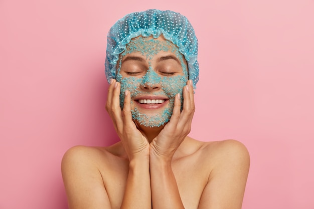 Skin care routine concept. Beautiful satisfied woman touches cheeks gently, applies nourishing sea salt facial scrub, satisfied by spa therapy, smiles broadly, wears blue waterproof headgear