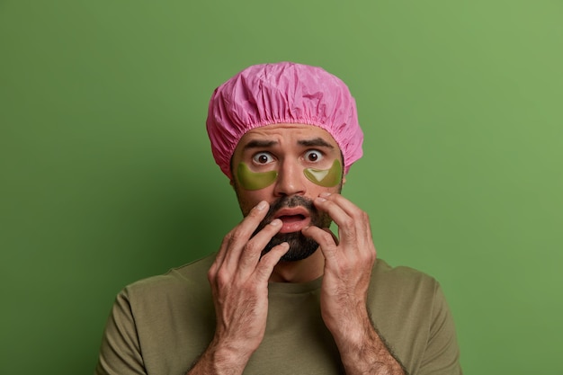 Free photo skin care routine, facial treatment. scared european guy wears collagen patches under eyes, cares about face, wears waterproof shower cap dressed in casual t shirt isolated on bright vivid wall.