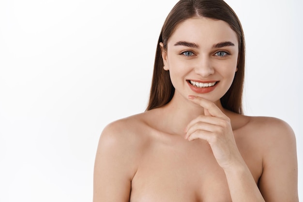 Skin care and spa. Beauty woman with clean smooth skin, blue eyes and perfect white smile, hold hand on chin, looking natural without make up, bare shoulders and moisturized glowing skin