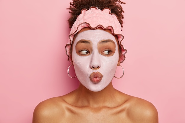 Skincare routine. Beautiful Afro American lady keeps lips rounded, applies nourishing mask on face, reduces chances of getting acne, gets little pleasure in life from beauty treatments, improves skin