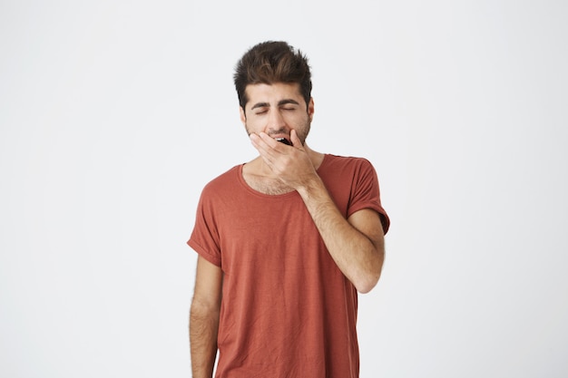 Sleepy beautiful caucasian man wearing red t-shirt covering mouth while yawning, after boring lecture at university. People and emotions concept