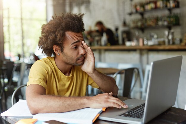 Sleepy college student with bushy hair and dark skin rubbing his eyes with hand while looking into screen of laptop wanting to sleep being tired preparing for final exams.