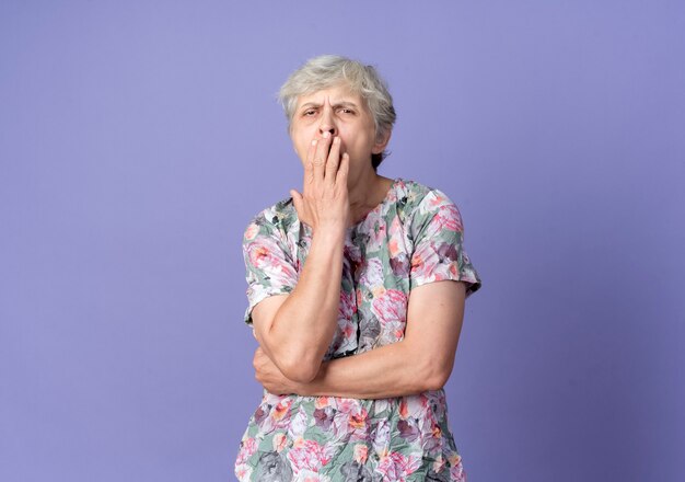 Sleepy elderly woman yawns and holds mouth isolated on purple wall