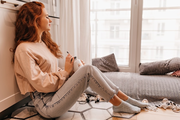 Free photo sleepy girl in jeans drinking mulled wine in cold day. indoor photo of curly young woman posing with cup of tea.