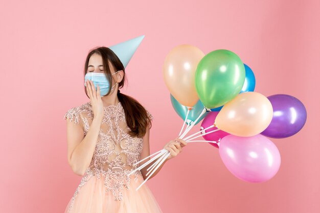 sleepy girl with party cap and medical mask holding colorful balloons on pink