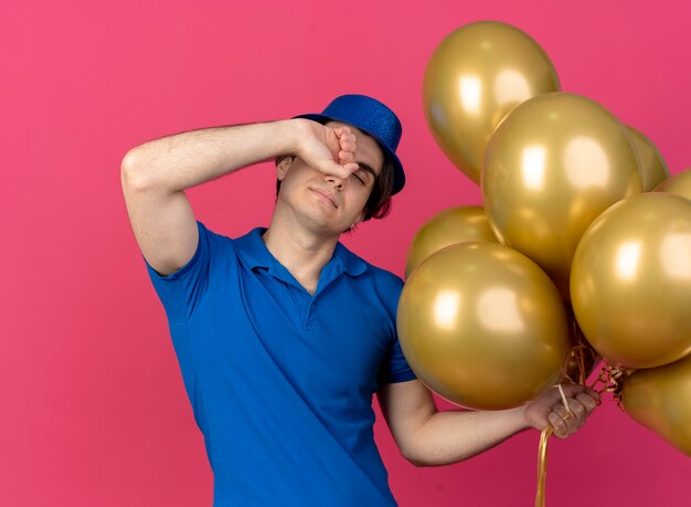 Sleepy handsome caucasian man wearing blue party hat puts fist on forehead and holds helium balloons 