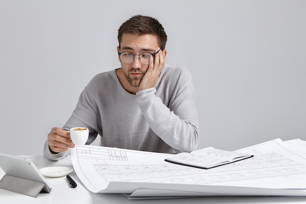 Free photo sleepy man drinks coffee as feels tired, works all day at blueprints, has fatigue expression