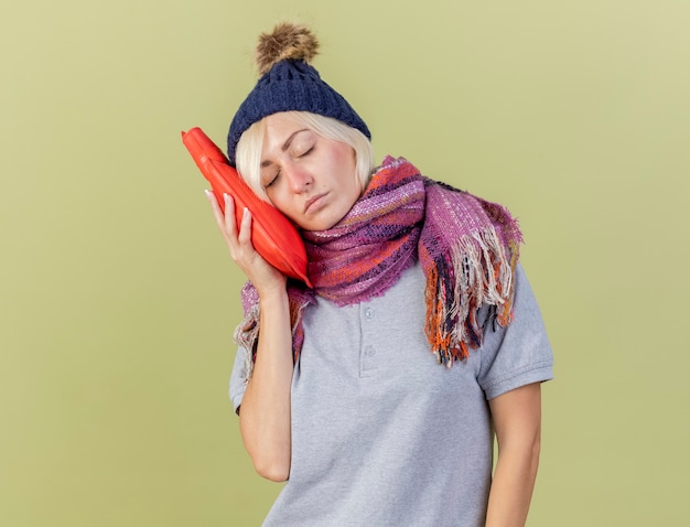 Free Photo sleepy young blonde ill slavic woman wearing winter hat and scarf puts head on hot water bottle isolated on olive green wall with copy space