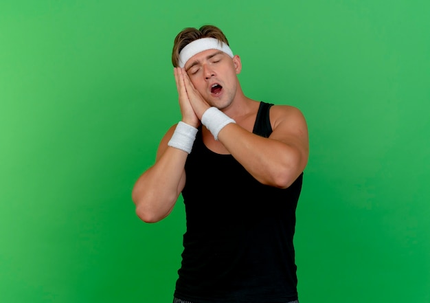 Sleepy young handsome sporty man wearing headband and wristbands doing sleep gesture with closed eyes isolated on green wall