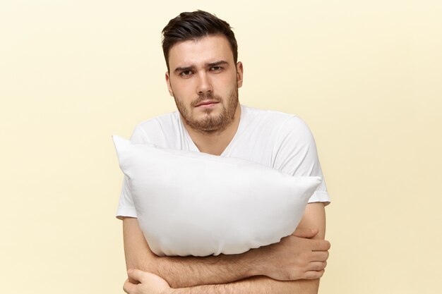 Sleepy young male posing with cushion. Attractive guy with bristle having tired facial expression because of insomnia trying to fall asleep, holding pillow.