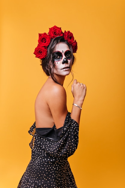 Slender dark-haired girl with beautiful posture posing in dress, baring shoulders. Portrait of mysterious Mexican lady with zombie masquerade mask