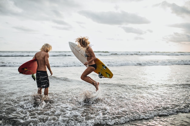 Slender guy and girl jump in sea water and hold surfboards