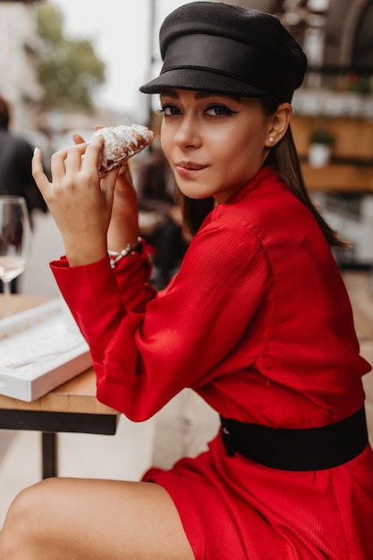 Free photo slender model could not restrain herself in front of delicious sweet croissant, sitting in parisian cafe. portrait of young lady in red outdoors