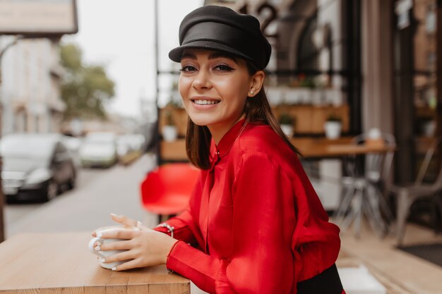 Slender woman in satin shirt and hat gently looks into lens. Brown hair lady enjoying her drink