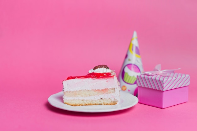 Slice of cake on plate with party hat and gift box on pink background