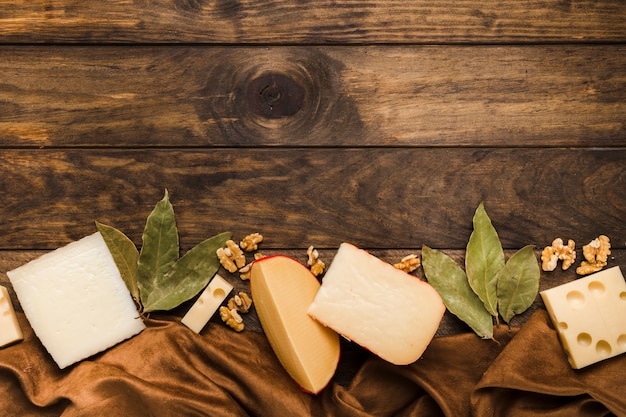 Slice of cheese; bay leaves and walnut arrange at bottom of the wooden background with silk material textile