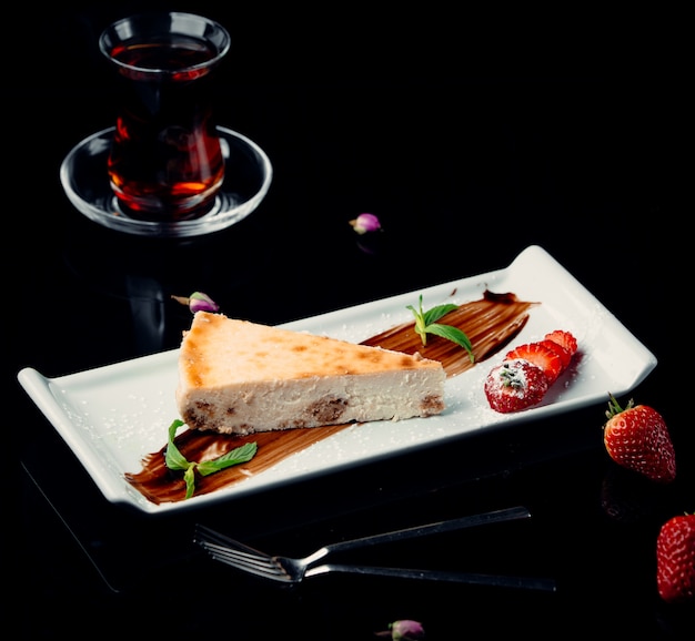 A slice of cheesecake with chocolate sauce,mint,strawberries and a glass of black tea.