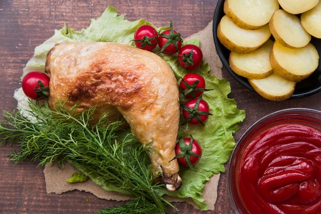 Free photo sliced boiled potato; roasted chicken leg; sauce; cherry tomato; dill for dinner on lettuce leaf over wooden table