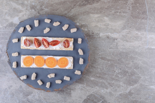 Sliced carrots, and tomatoes on crispbreads, surrounded by crumb on the board, on the marble surface