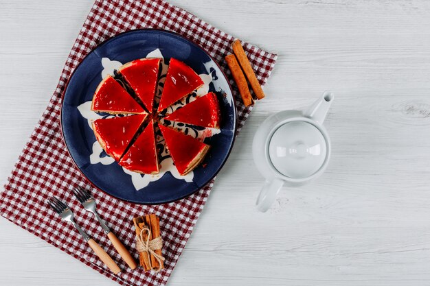 Sliced cheesecake in a plate and picnic cloth with a teapot, two forks and cinnamon sticks flat lay on a light grey background