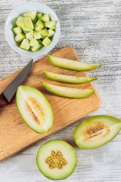 Sliced melon in a wooden cutting board with melon in bowl and knife flat lay on a white stone background